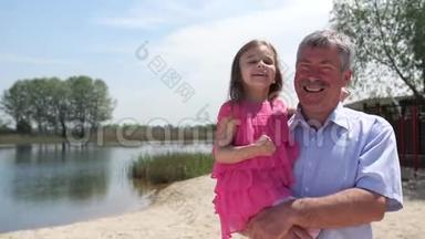 祖父和孙女沿着海滩散步。 那个女孩和她的祖父坐在他的手上。 他们玩得很开心