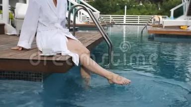 优雅的女人穿着<strong>白色</strong>浴袍，坐在豪华<strong>别墅</strong>的泳池边的码头上，在泳池里着脚。