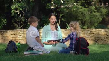 年轻的女教师在户外为小男孩和小女孩上课，手里拿着书，坐在草地上