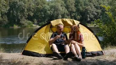 心满意足的幸福夫妇坐在帐篷里。 旅游和旅游的概念.. 女孩用热水瓶倒茶. 森林和河流