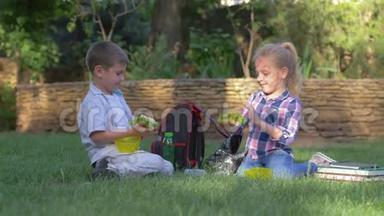 孩子们在课间休息时吃午餐，坐在背包旁边，在草坪上看书，男孩和女孩在户外吃三明治