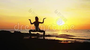 年轻女子`她的剪影正在日落时在海边做瑜伽练习。