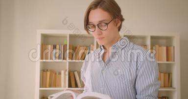 一位年轻成功的高加索男学生戴着眼镜看着图书馆里的照相机看书的特写肖像