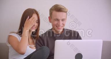 一对年轻可爱的白种人夫妇用笔记本电脑一<strong>起坐</strong>在室内沙发上看电影的特写镜头