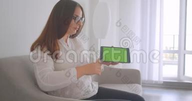 特写侧景照片，年轻的白种人妇女戴着眼镜，使用平板电脑，并显示绿色的彩色屏幕，以显示。
