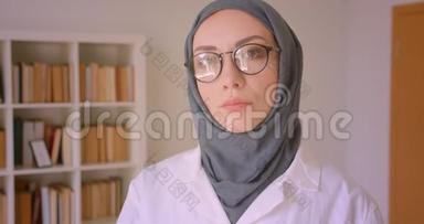 阿拉伯青年女医生戴着眼镜和头巾，在图书馆里兴高采烈地看着镜头的特写照片