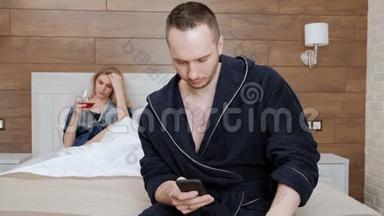 一对年轻的已婚夫妇穿着浴袍躺在酒店房间卧室的一张白色床上。 一个男人在