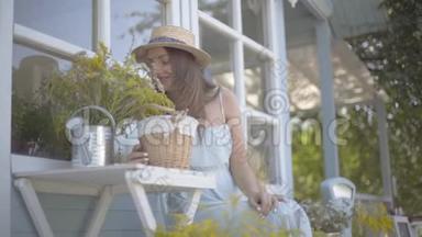 戴着草帽和白色裙子的迷人年轻女子微笑着，坐在浇花的罐子里嗅着野花