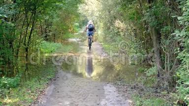 一个女人骑着自行车走在泥路上，<strong>身边</strong>有水坑