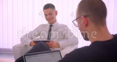 在办公室里用笔记本电脑向他的伴侣展示带有绿色屏幕的平板电脑的快乐商人的特写肖像