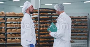 年轻的面包师和主要工程师在一个面包店区进行了一次谈话，那里有很多货架上有新鲜的烘焙面包
