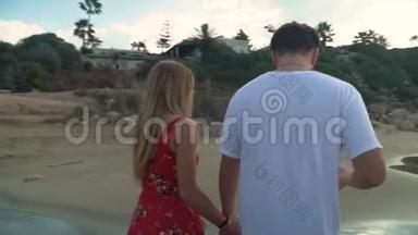 可爱的一对夫妇沿着海滨散步，紧紧地牵着手。 穿白色t恤的高个子男人和穿红色衣服的女人休息