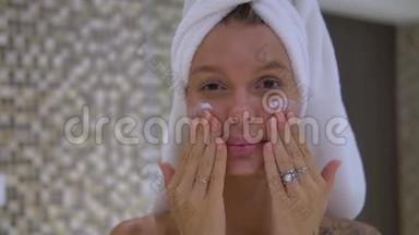 一位<strong>身材矮小</strong>可爱的年轻女士，头上戴着白色毛巾，早上在浴室里用化妆品按摩脸部