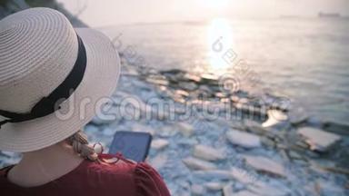 从后面可以看到一个漂亮的年轻女孩，她穿着一件夏天的红色连衣裙和一顶草帽，坐在海边的石头上