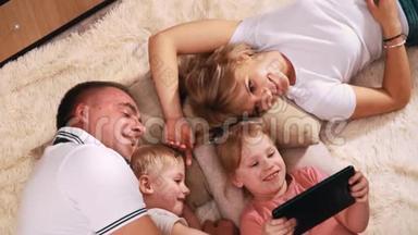 一家人带着<strong>两个小孩子</strong>躺在温暖的毯子上，微笑着看着平板电脑，它抱着她的女儿。 爸爸
