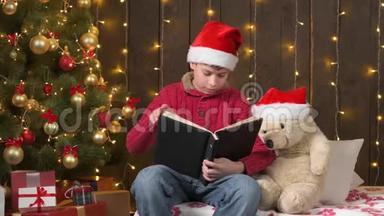 圣诞老人帮手男孩看书熊玩具，坐在室内靠近装饰圣诞树灯-圣诞快乐和节日快乐