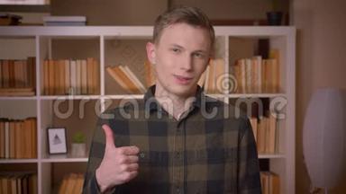 一位年轻迷人的白种人男学生的特写镜头，他竖起大拇指看着大学图书馆里的摄像机