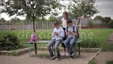 两个男孩和女孩在放学后使用他们的电话。 可爱的男孩坐在长凳上玩网络游戏。 女孩的样子