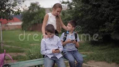 两个男孩和女孩在<strong>放学</strong>后使用他们的电话。 可爱的男孩坐在长凳上玩网络游戏。 女孩的样子