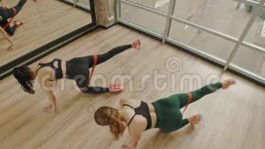 两个女人在工作室里用大腿之间的拉伸皮带训练腿-站在膝盖上，拉着腿