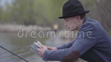 一位穿着夹克衫、戴着野帽的时尚渔夫坐在河边，看着一本书或钓鱼提示