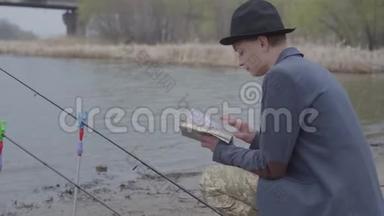 一位穿着夹克衫、戴着野帽的时尚渔夫坐在河边，看着一本书或钓鱼提示