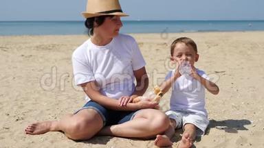 在阳光明媚的夏日，妈妈和儿子正坐在海滩上。 家庭度假和自然野餐。 孩子吃饭，吃饭