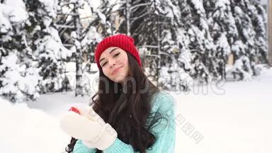 年轻漂亮的女孩穿着一件温暖的蓝色毛衣，戴着手套，背景是白雪覆盖的树木，抱着一颗红色的心，