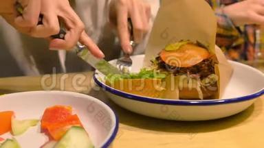 女人正在吃大汉堡，用叉子和刀在盘子上端着，双手特写。