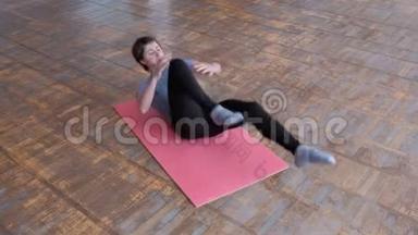 穿着灰色T恤和黑色紧身衣的运动女孩躺在粉红色的地毯上，两腿交替弯曲以加强腹部