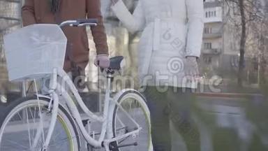 无法辨认的女人穿着温暖的白色夹克，高个子男人穿着棕色外套，骑着自行车走在街上。 休闲