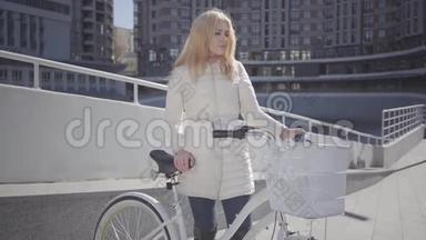 美丽的金发微笑的女人，穿着温暖的白色夹克，站在城市街道上，骑着自行车看着镜头。 休闲