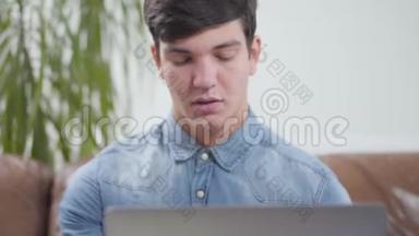 肖像英俊的年轻人坐在家里的笔记本电脑前。 学生检查邮箱。 对小玩意的上瘾