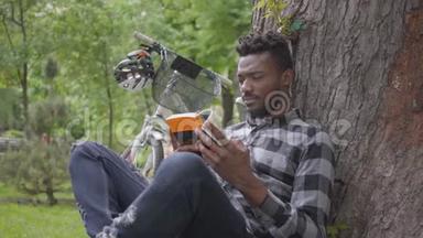 一位英俊、自信的非洲裔美国人坐在公园的一棵老树下的自行车旁看书