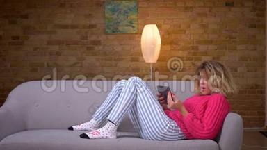 金发的家庭主妇，穿着粉红色毛衣，躺在沙发上，在舒适的家庭氛围中，在平板电脑上视频聊天。
