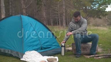 一个年轻人坐在森林里蓝色帐篷附近的一根大木头上，从热水瓶里倒茶。