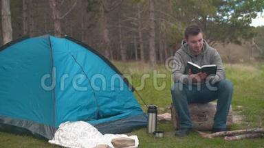 一个年轻人坐在一个大圆木附近的蓝色帐篷和Temos在森林里读一本书。