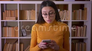 美丽的黑发老师戴着眼镜看着智能手机，<strong>聚精会神</strong>地站在图书馆。