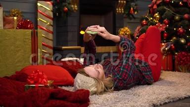 年轻漂亮的女孩或女人穿着红色格子衬衫躺在地板上，拿着圣诞树通过电话进行视频通话