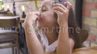 肖像小女孩吃甜蛋糕，在咖啡馆的桌子上微笑。 面对可爱的女孩微笑着吃着甜馅饼