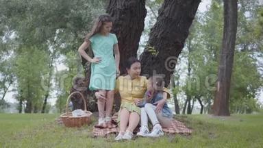 优雅的高级女人坐在公园树下的毯子上和两个可爱的孙女交谈。 一个女孩