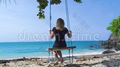 穿着黑色裙子的年轻女子在海滩上摇摆着蓝色的海洋景观。 快乐的女人在夏日海滩上荡秋千