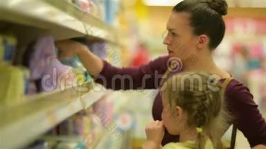 可爱的女孩和棒棒糖坐在超市手推车。 母亲在后台选择湿巾。