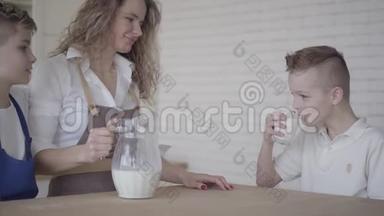 一个男孩在厨房里坐在他母亲和哥哥旁边的桌子旁喝牛奶。 兄弟情谊。 家庭