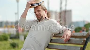 放松迷人的中年男子，长发，戴着帽子，坐在公园的长凳上休息。 夏季傍晚，炎热..
