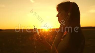 女孩在日落的光线中旅行和听音乐和舞蹈。 戴着耳机和智能手机的年轻女孩