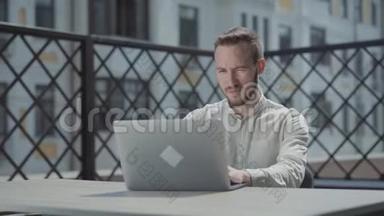 肖像自信胡须的年轻人坐在笔记本电脑前的露台上的桌子旁工作。 概念
