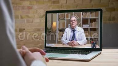 戴着白色实验室外套和眼镜的资深医生在网上视频卡上与屏幕后的病人认真交谈。