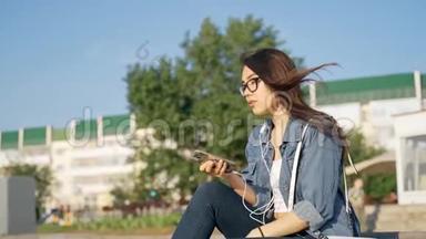 一个戴眼镜的年轻女孩坐在街上，用耳机听音乐。 手里拿着手机的女孩