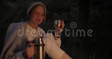 一个年轻的女人晚上坐在火对面的森林里，从<strong>热水</strong>瓶里倒茶<strong>喝</strong>。